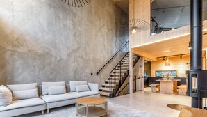 Una sala de estar moderna con un sofá de color claro, una mesa redonda de madera y una escalera que sube a un nivel superior.