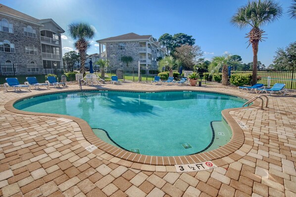 A swimming pool with blue water, surrounded by lounge chairs and palm trees. - Coastal Fairways Golf Course Condo 5 Min to Beach (Myrtle Beach)