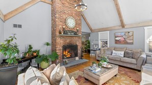 A spacious living room with a brick fireplace, a large sofa, and multiple potted plants. - Barnstable Cape Cod Retreat | Pool, Fire Pit & BeachSticker (Barnstable)
