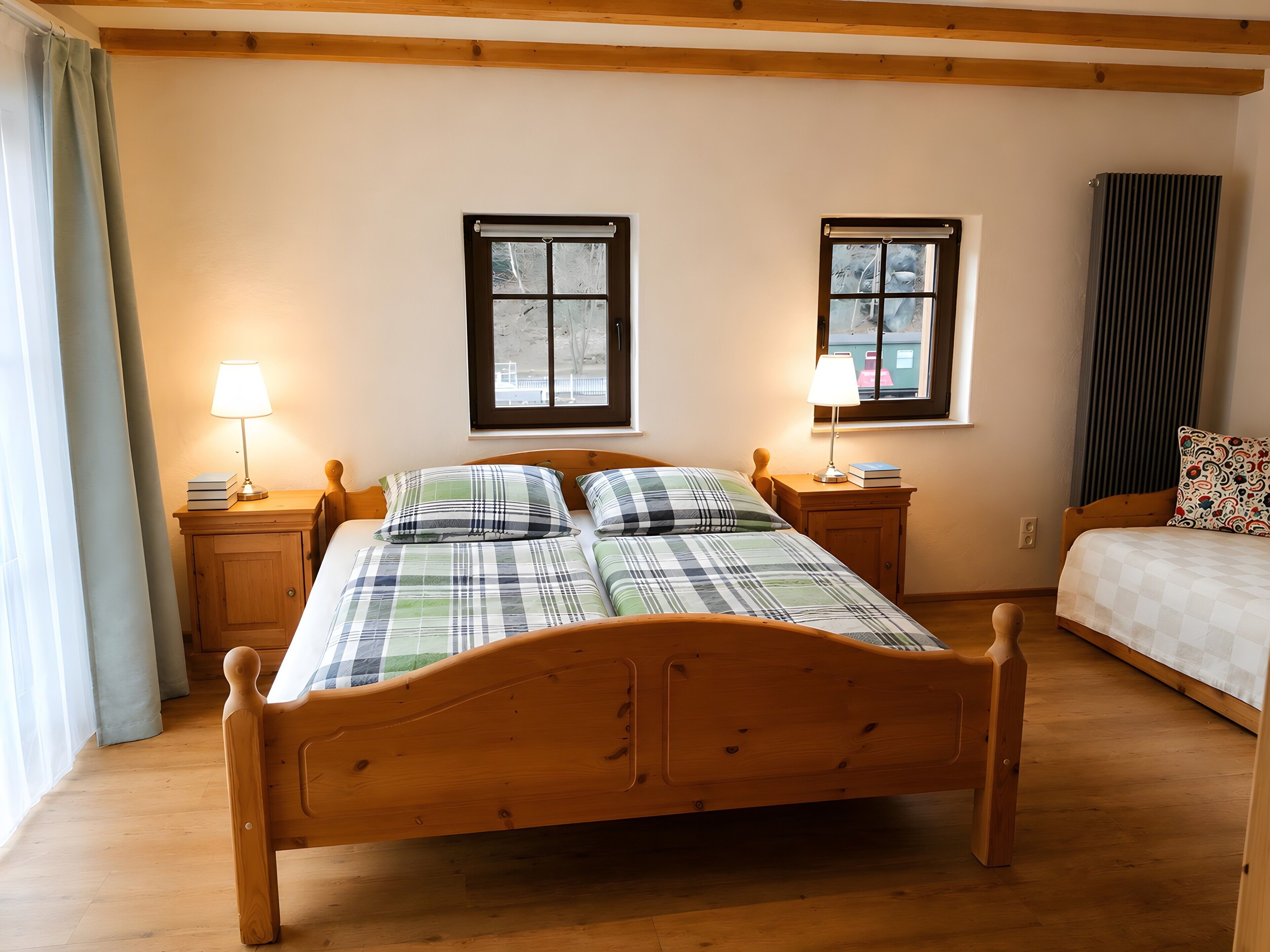 A wooden bed in a room with two windows, a bedside table with a lamp, and a radiator.