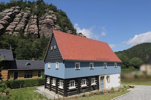 A traditional house with a red roof, white window frames, and a blue facade, surrounded by a garden and a rocky mountain backdrop.