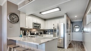 A modern kitchen with white cabinets, a central island, bar stools, a microwave, and a refrigerator. - ⛱️Oceanfront | Two Screened Balconies | 1st Floor Unit | Tilghman Beach 127🌊 (North Myrtle Beach)