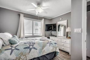 Un dormitorio con cama, ventilador de techo, ventana con persianas, cómoda y espejo.