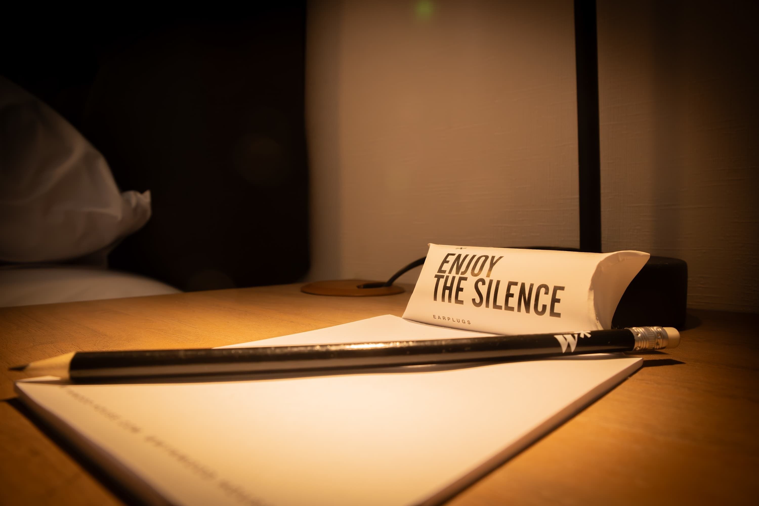 A desk with a notebook, a pen, and a sign that reads "ENJOY THE SILENCE" with earplugs underneath.