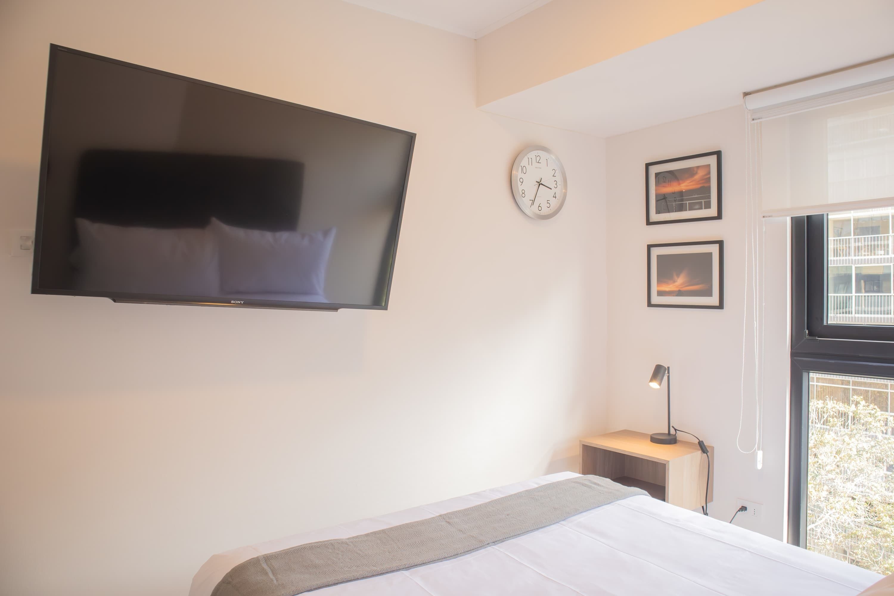 A bedroom with a flat-screen TV mounted on the wall, a bedside table with a lamp, a clock, and two framed pictures.
