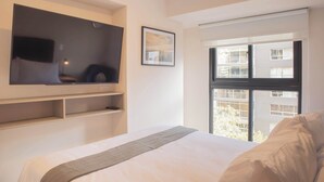 A hotel room with a bed, a mounted TV, a window with a view of buildings, and a framed picture on the wall. - w Cozy 2BR w Balcony in Miraflores (Lima)