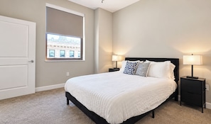 Une chambre à coucher avec un grand lit, deux tables de chevet avec des lampes, une fenêtre donnant sur des immeubles et une porte.