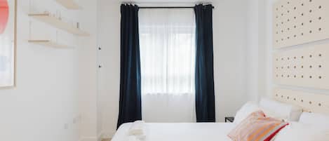 Une chambre d’hôtel comprenant un lit, une table de chevet, une étagère fixée au mur, une fenêtre avec des rideaux et une tête de lit ornée d’un motif.