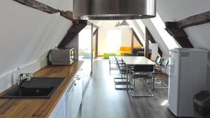 A compact kitchen with a central island, a dining area with a table and chairs, and a high ceiling with a large vent. - Les Chambres de Louise (Habsheim)