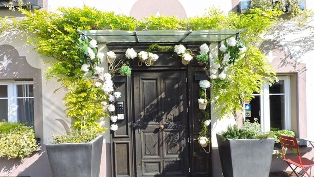 A black door with a white floral archway, flanked by large planters and a red folding chair. - Les Chambres de Louise (Habsheim)
