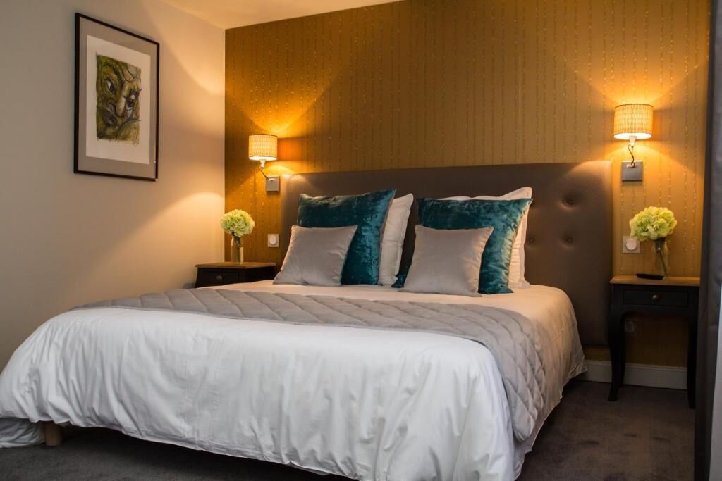 Una cama cuidadosamente tendida con cubrecama blanco y almohadas turquesa, flanqueada por dos mesitas de noche con lámparas y pequeños jarrones con flores.