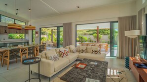 A spacious living area with a large sofa, dining table, and chairs, a coffee table, and a kitchen area with a view of greenery. - Eko Savannah by MJ Holidays (Tamarin)