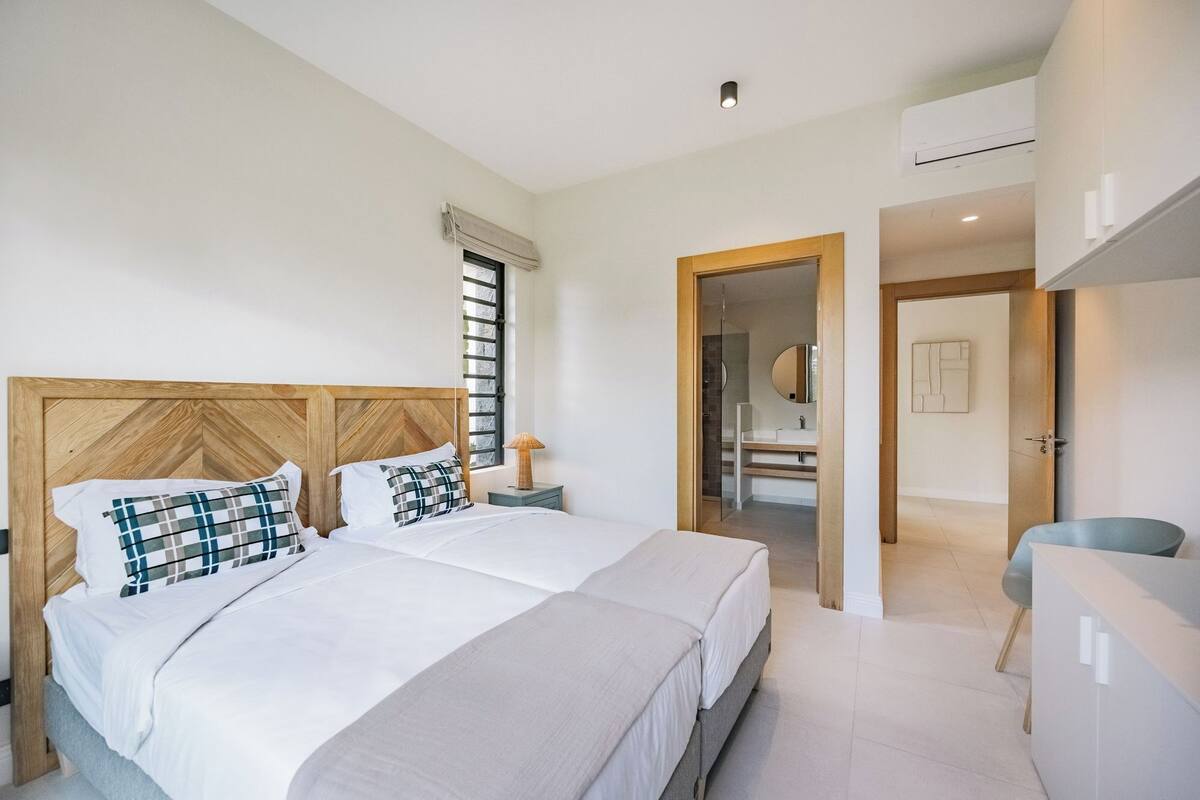Ein modernes Hotelzimmer mit zwei Betten, einem hölzernen Kopfteil, einem Fenster mit Jalousien, einem Nachttisch mit Lampe und einem durchgegangenen Tür angedeuteten Badezimmer.