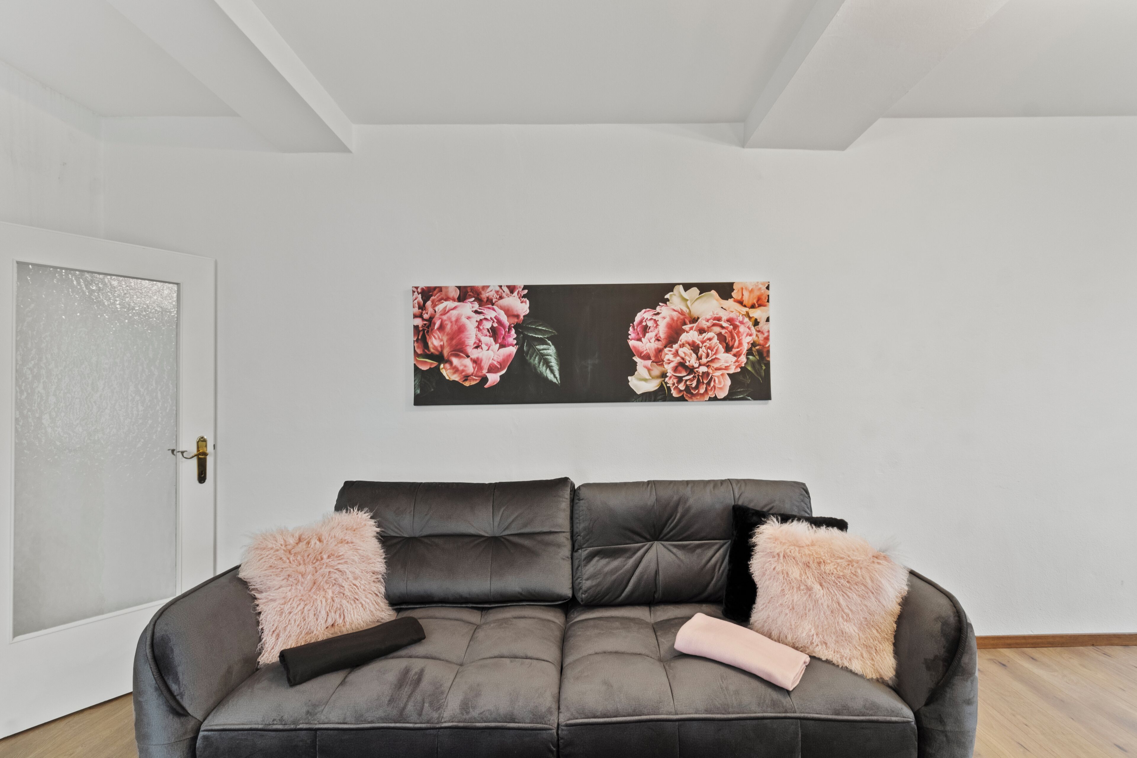 A modern living room with a dark grey sofa, two fluffy cushions, and a large floral painting on the wall.