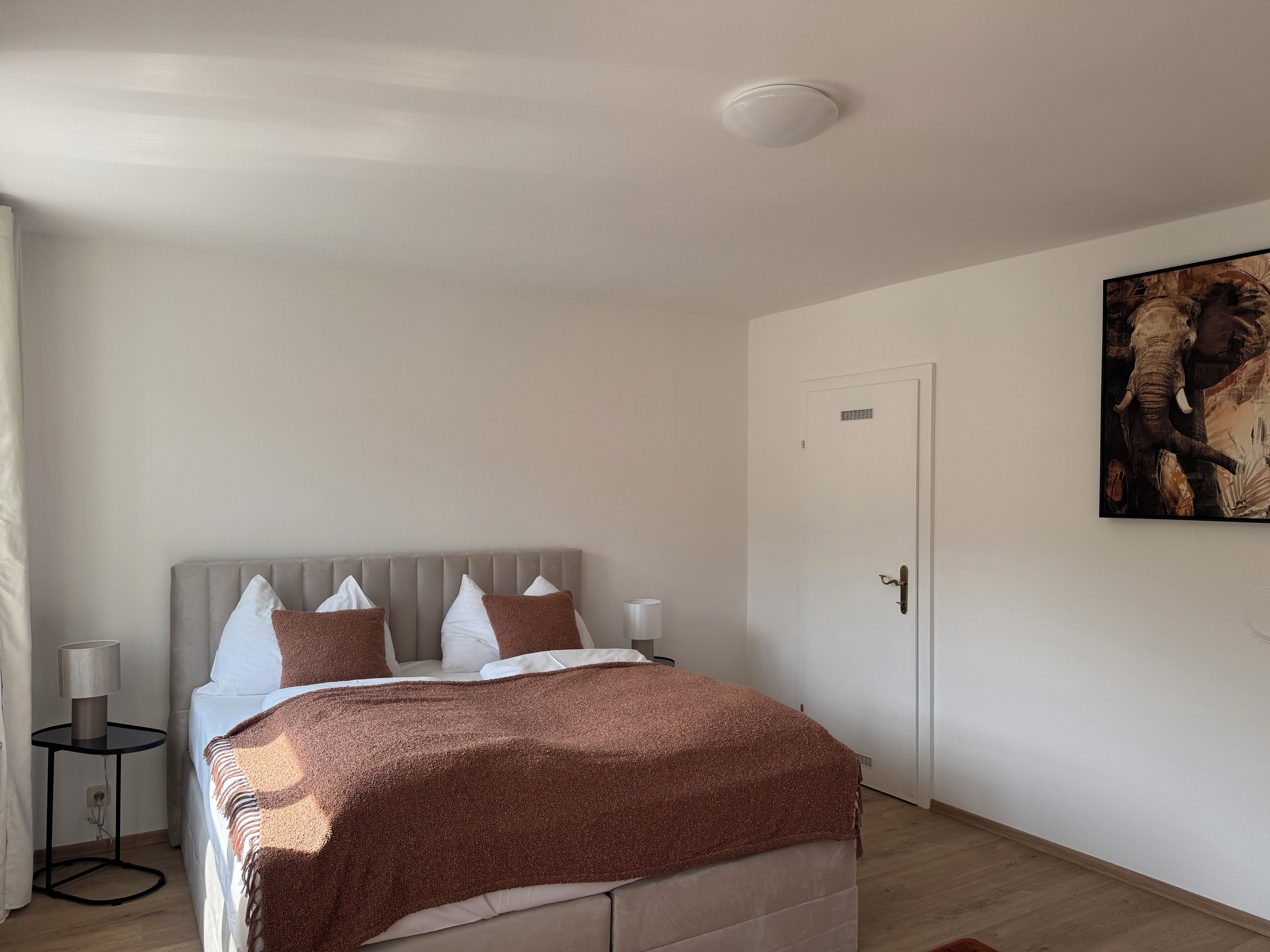 Un dormitorio con una cama, una mesita de noche, una lámpara, una puerta y un cuadro en la pared.