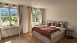 Une chambre à coucher avec un grand lit, deux fenêtres avec des rideaux, un radiateur et une table de chevet avec une lampe.
