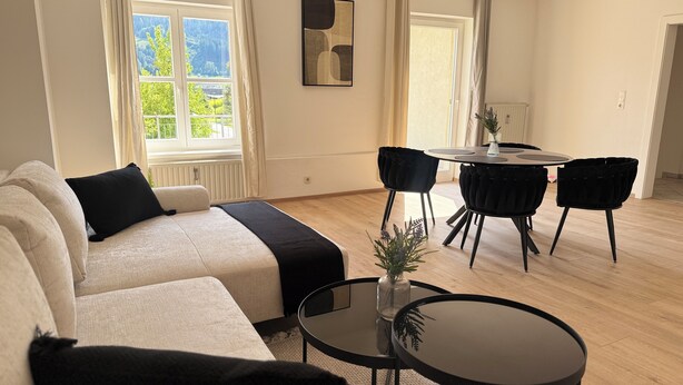 Un salon moderne avec un canapé, deux fauteuils noirs, une petite table et une vue sur un paysage verdoyant par la fenêtre.