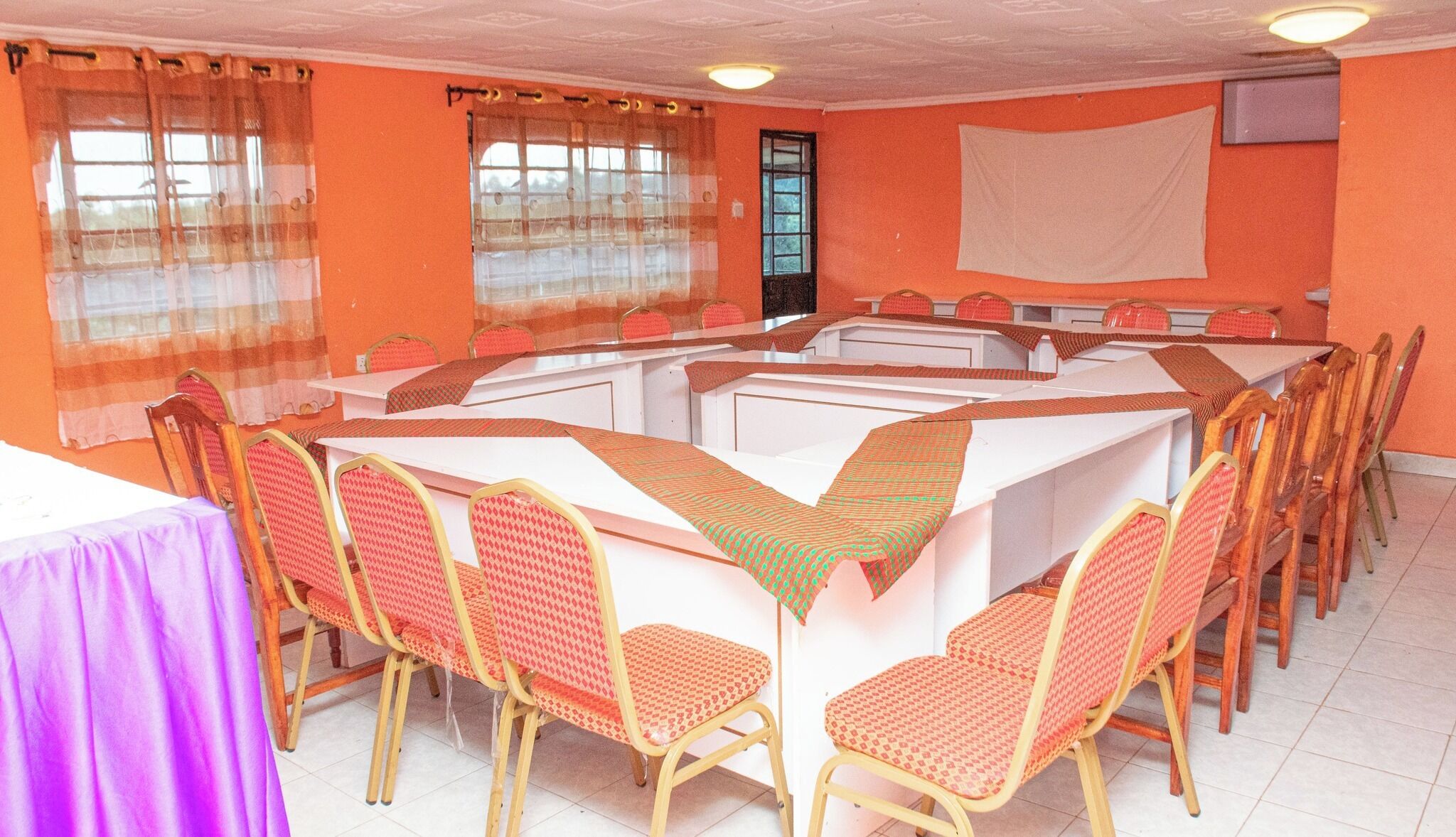 Une salle de conférence avec une table centrale entourée de chaises, des murs orange et de grandes fenêtres ornées de rideaux légers.