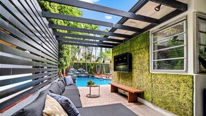 A covered outdoor seating area with a pool, a TV mounted on the wall, and a wooden bench. - Beach Pop - Steps to Beach, Heated Pool & Hot Tub (Fort Lauderdale)