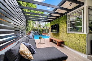 A covered outdoor seating area with a pool, a TV mounted on the wall, and a wooden bench. - Beach Pop - Steps to Beach, Heated Pool & Hot Tub (Fort Lauderdale)