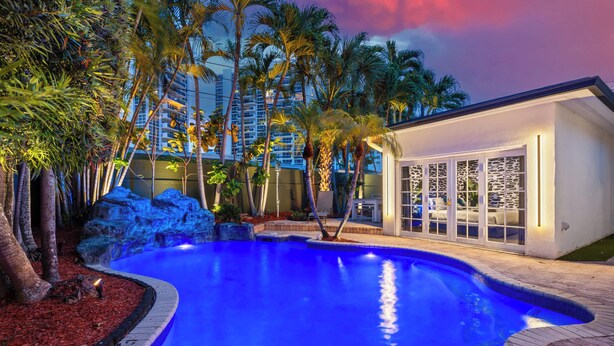 A swimming pool with a waterfall feature, surrounded by palm trees and a modern building with large windows. - Beach Pop - Steps to Beach, Heated Pool & Hot Tub (Fort Lauderdale)
