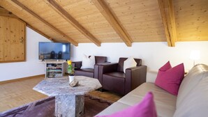 Un salon chaleureux avec une table basse en pierre, des canapés en cuir et un téléviseur à écran plat fixé à une poutre en bois au plafond.