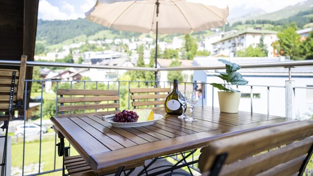 Une table en bois, une bouteille de vin, deux verres, une assiette de raisins et une plante en pot, le tout sur un balcon abrité par un parasol.