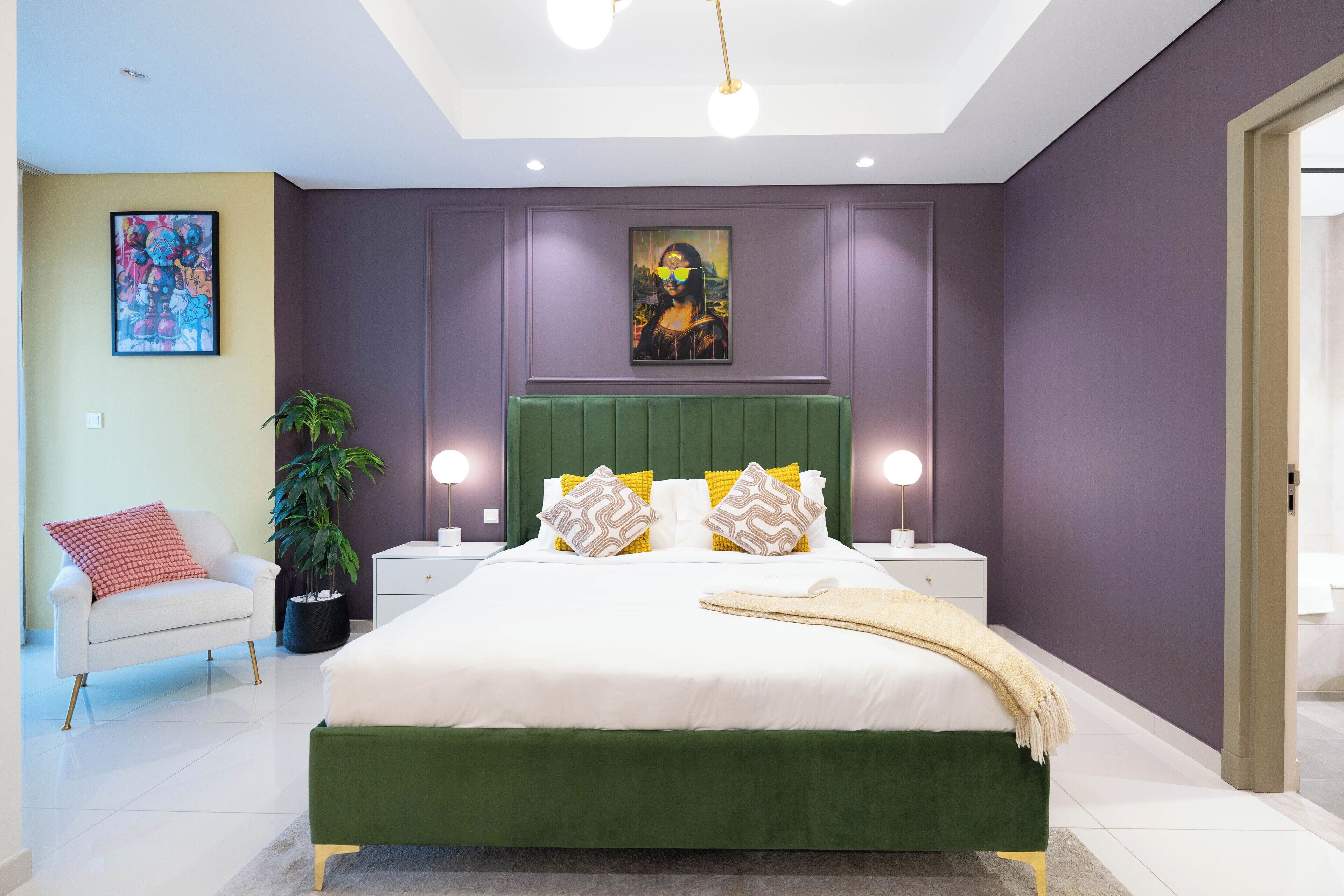 Une chambre moderne avec un lit recouvert d’un tissu vert, du linge de lit blanc et des coussins décoratifs. On y trouve une table de chevet avec une lampe, une plante en pot et une œuvre d’art encadrée au-dessus du lit.