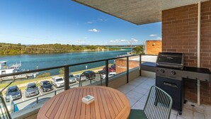 A balcony with a view of a river, a barbecue grill, a round wooden table, and a parking area with cars. - Lago Vista on the Forster lakefront (Forster)