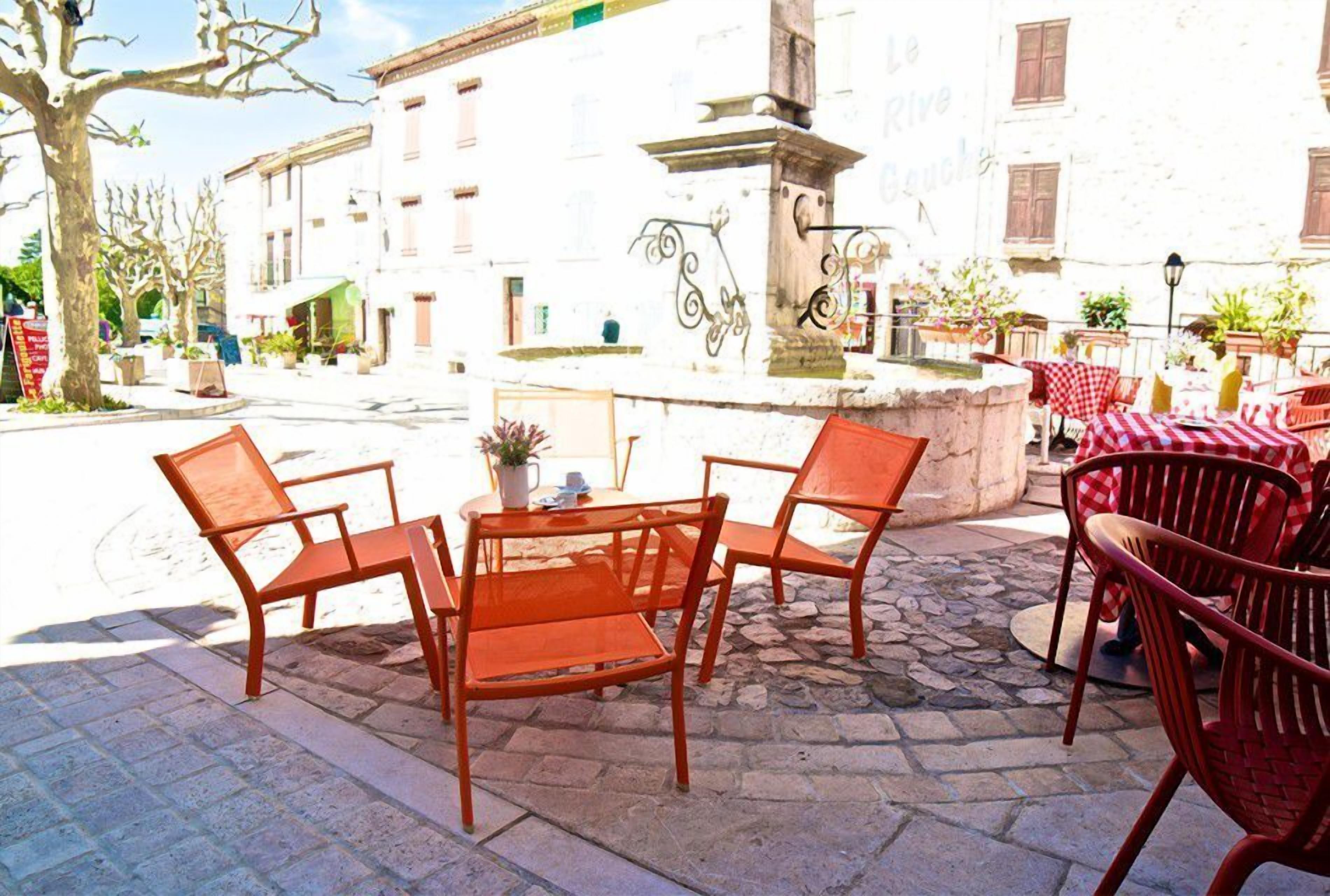 Un espace extérieur aménagé avec des chaises et des tables en métal orange, une fontaine en pierre et, au fond, un bâtiment à la façade blanche et aux volets rouges.
