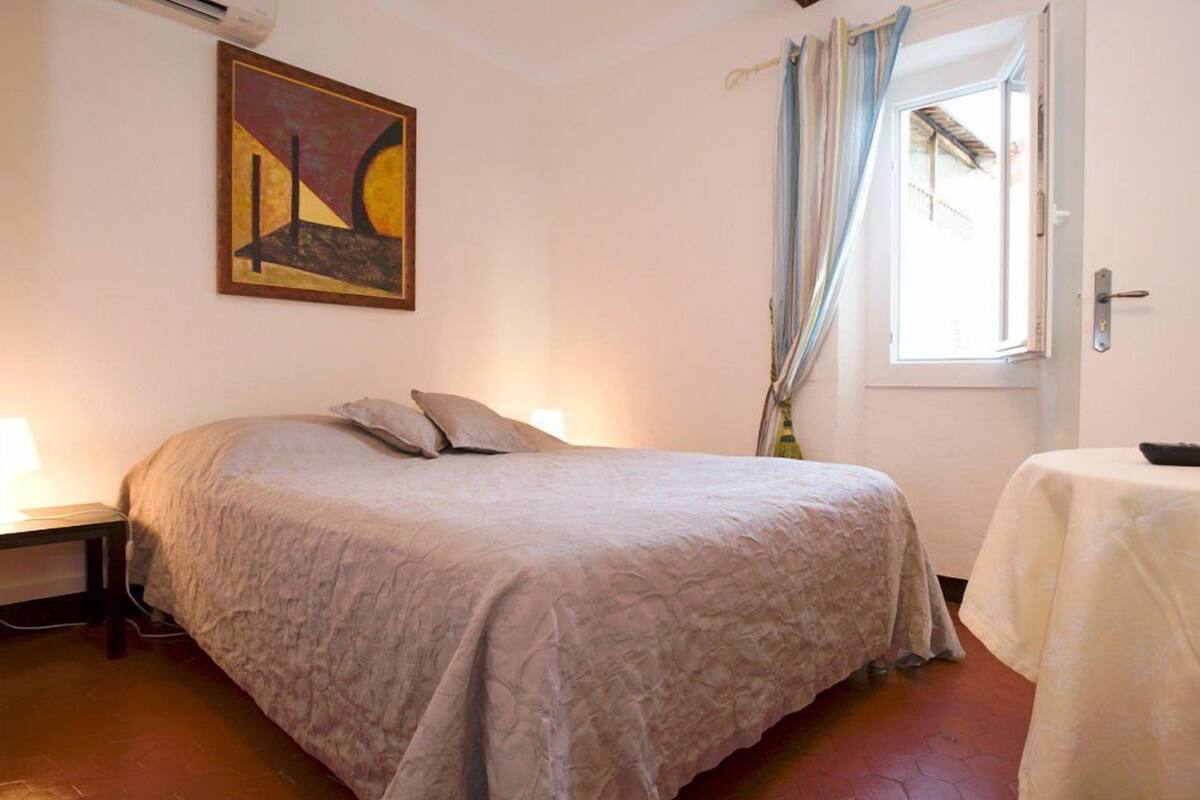 Un dormitorio con cama, mesita de noche, una mesa, ventana con cortinas y un cuadro en la pared.