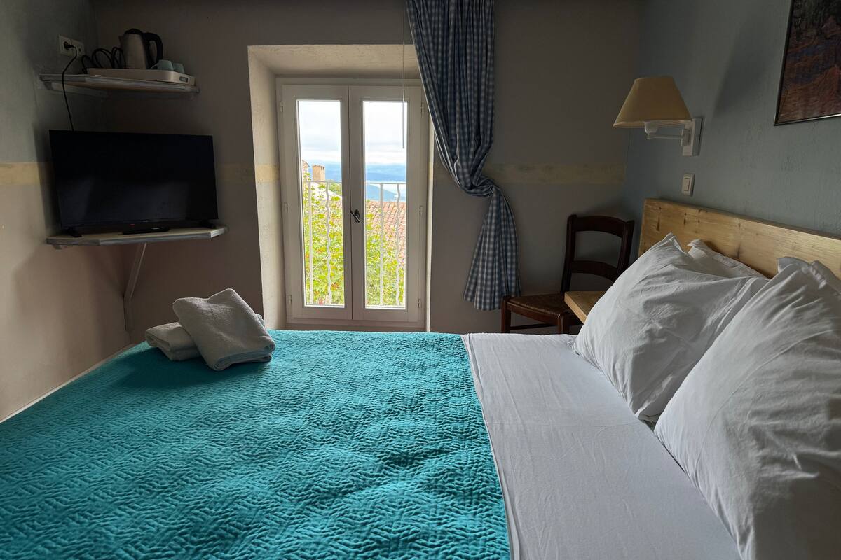 Habitación de hotel con cama, televisor en una estantería, una silla y vistas al exterior a través de una puerta.