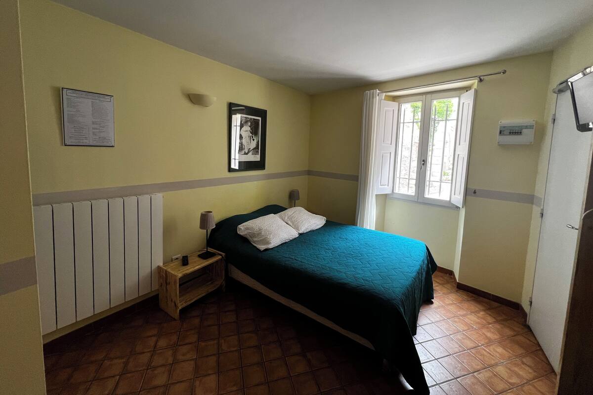 Un dormitorio con cama, mesita de noche, ventana con cortinas, radiador y un cuadro eléctrico en la pared.