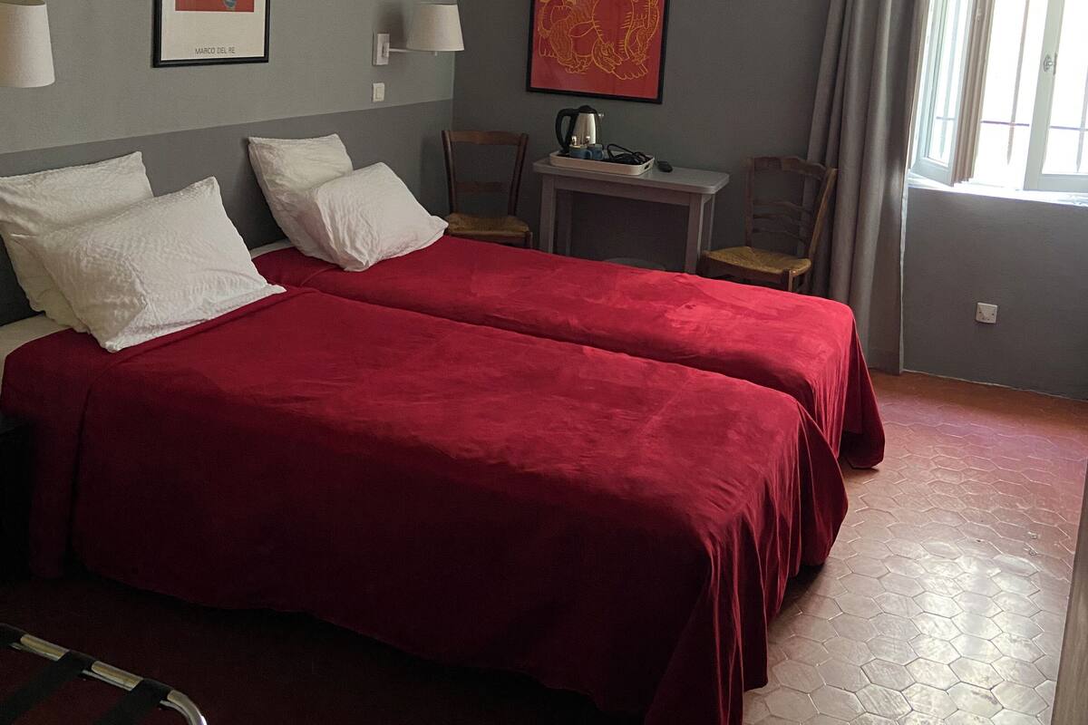 Habitación de hotel con dos camas, una mesita con un hervidor, una silla y dos cuadros enmarcados en las paredes.