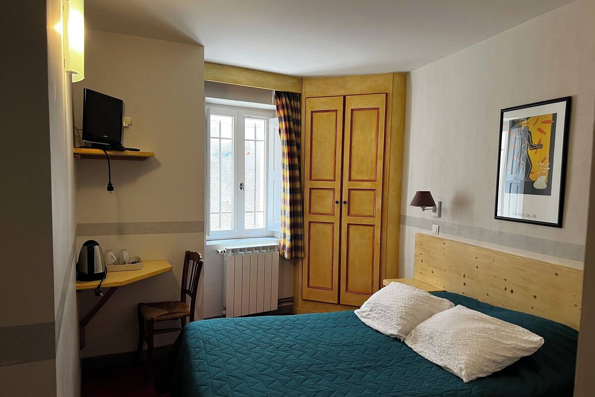 Un dormitorio con cama, una silla, un escritorio, ventana con cortinas, radiador, televisor sobre una estantería y un cuadro en la pared.