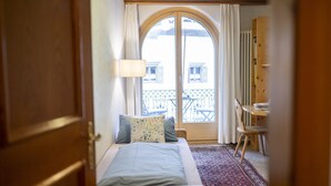 A bedroom with a bed, a desk, a chair, a bookshelf, and a view of a balcony with outdoor furniture. - Chasa Bügliet: Holiday flat in the village center of Sent (Sent)