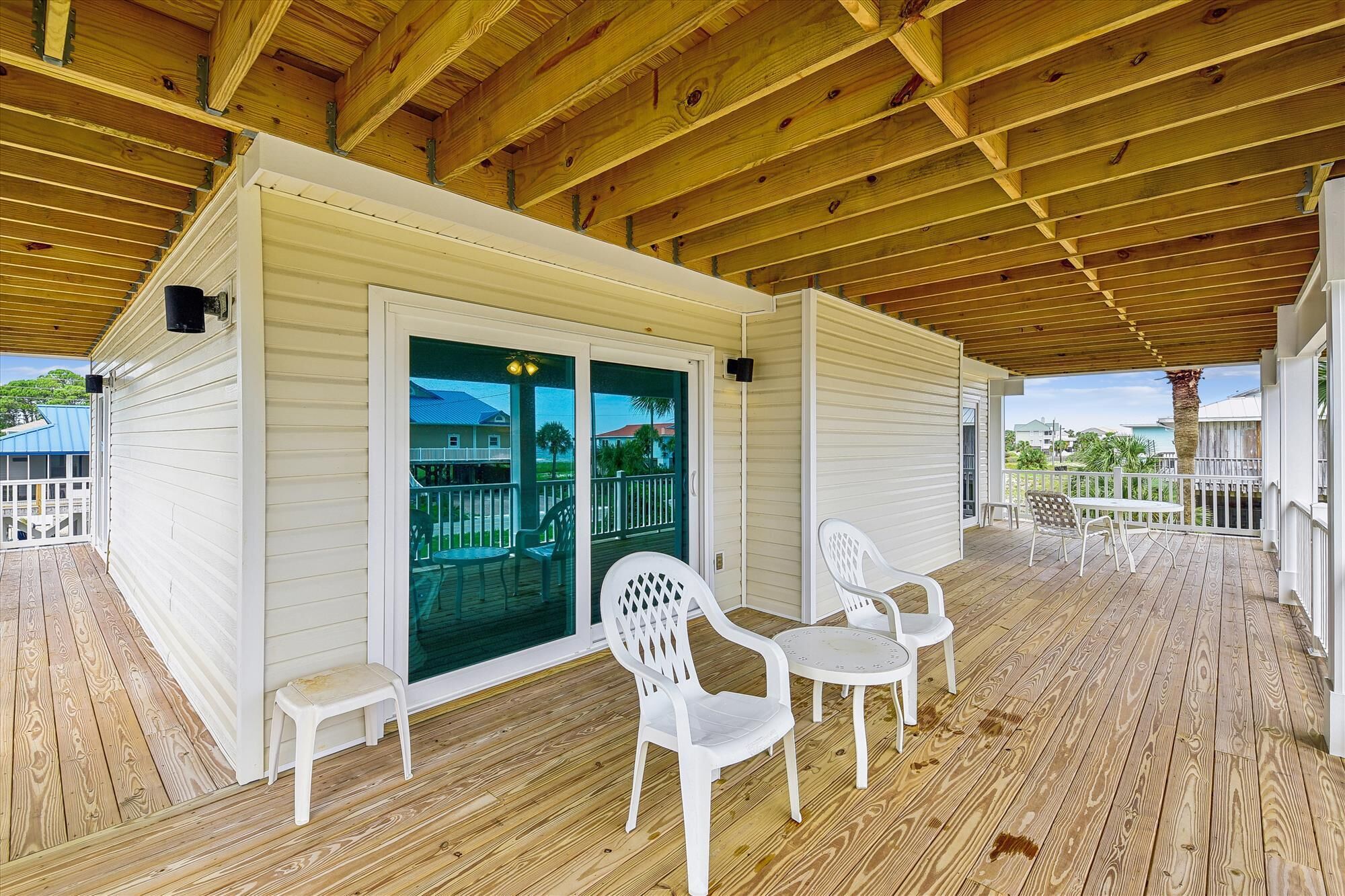 A covered porch with white plastic chairs and a small table, wooden flooring, and a view of the ocean and other buildings.