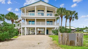 A two-story beachfront house with a covered porch, multiple balconies, and a gravel driveway.