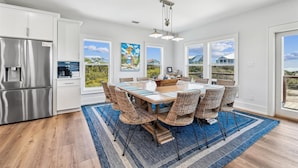 A modern kitchen with stainless steel appliances, a dining area with wicker chairs, and large windows offering views of the outdoors.