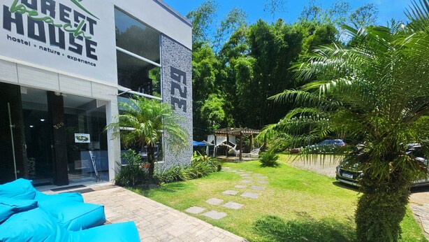 A modern hostel with a glass facade, outdoor seating area with blue cushions, and a well-maintained garden with palm trees. - Foresthouse Beach Hostel (Florianópolis)