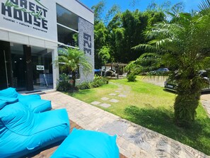 A modern hostel with a glass facade, outdoor seating area with blue cushions, and a well-maintained garden with palm trees. - Foresthouse Beach Hostel (Florianópolis)