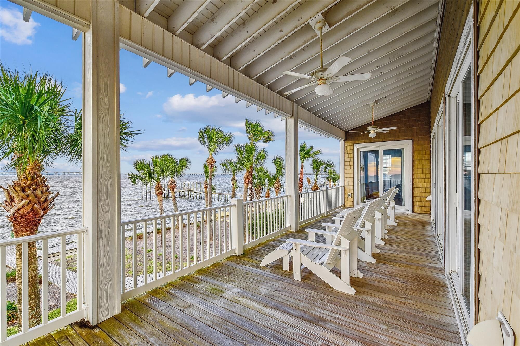 A covered wooden deck with white chairs, a railing, and a view of the sea with palm trees.