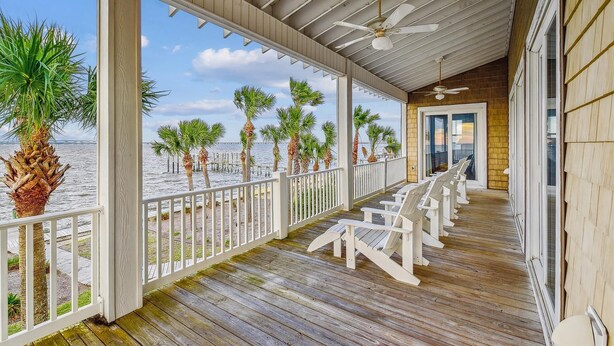 A covered wooden deck with white chairs, a railing, and a view of the sea with palm trees.