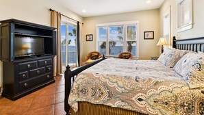 A bedroom with a large bed, a TV, a ceiling fan, and a view of the ocean through the window.
