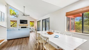 Un salon moderne avec un canapé blanc, une table à manger avec des chaises, une cheminée et une grande fenêtre donnant sur la forêt.