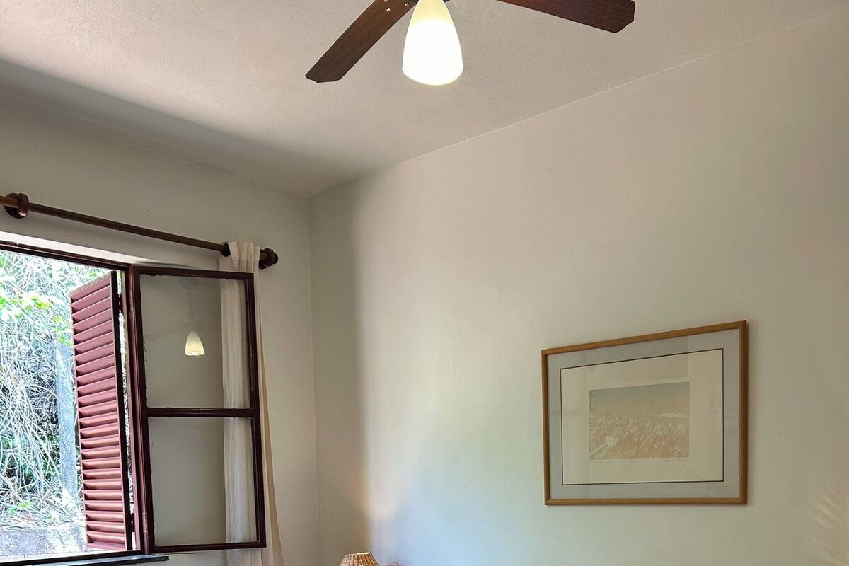 Un lit bien fait, avec du linge de lit blanc, une tête de lit en bois et un plaid marron. On y trouve une lampe de chevet, un tableau encadré au mur et une fenêtre avec des rideaux.