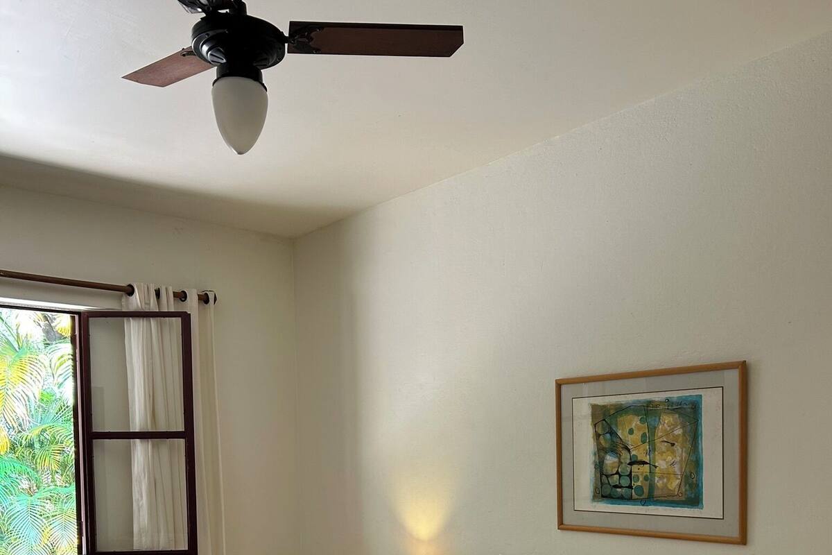 Une chambre d’hôtel avec deux lits, un ventilateur de plafond, une fenêtre avec des rideaux, une lampe de chevet et un tableau encadré au mur.