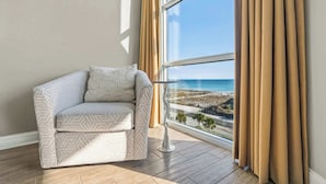 A corner room with a view of a beach and ocean through a large window, a corner sofa with patterned cushions, and a floor lamp.