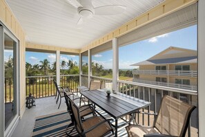 A covered outdoor area with a table and chairs, large windows, and a view of palm trees and buildings. - Surfside 12 A4 - Stunning, 3 bedroom, 2 bathroom condo with water views! (Sanibel Island)