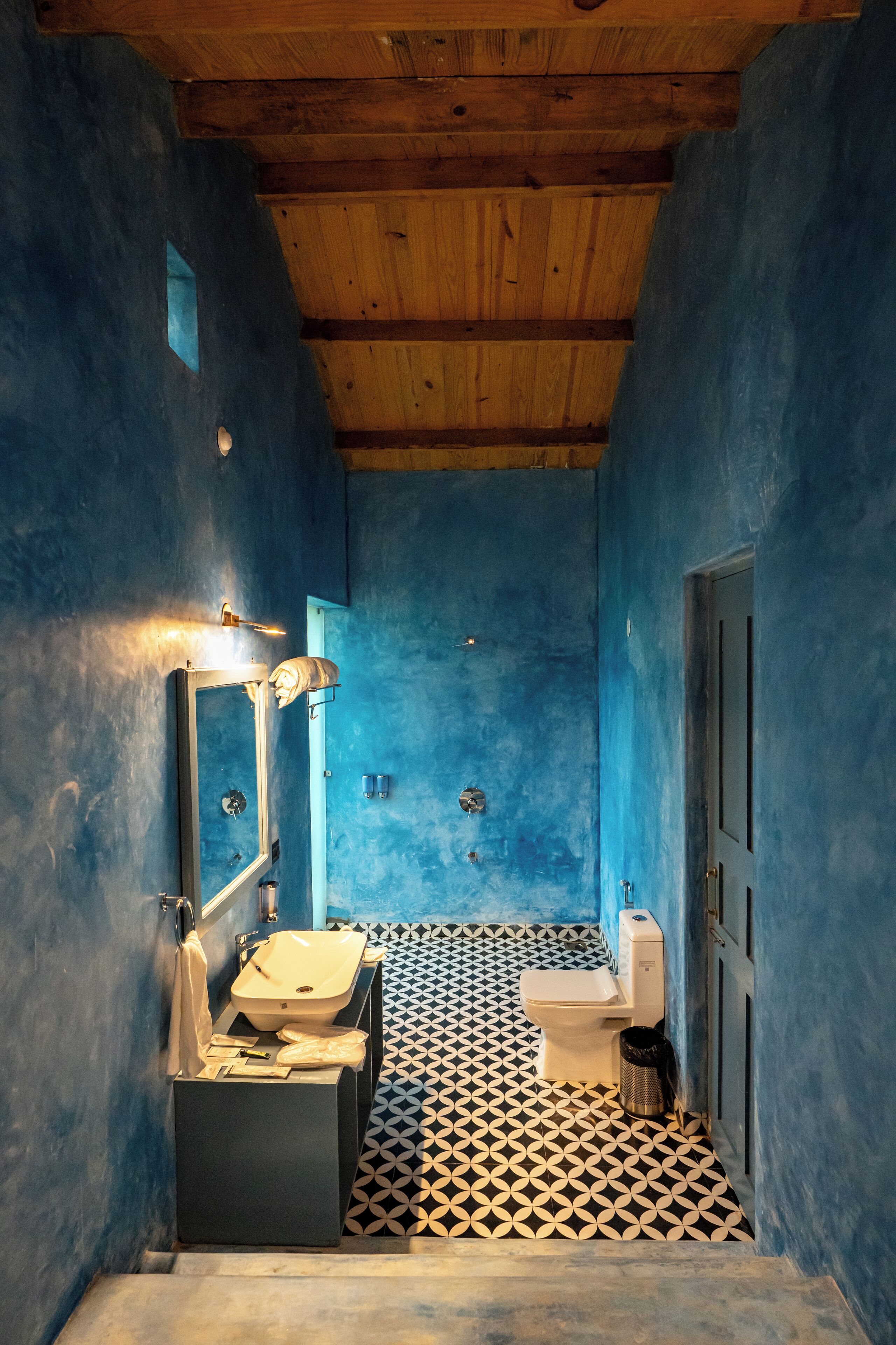 A narrow bathroom with blue walls, a wooden ceiling, a black and white patterned floor, a sink, a toilet, and a mirror.